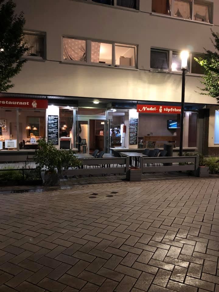 Restaurantfassade bei Nacht mit rotem Nudeltöpfchen-Schriftzug und Außenplätzen
